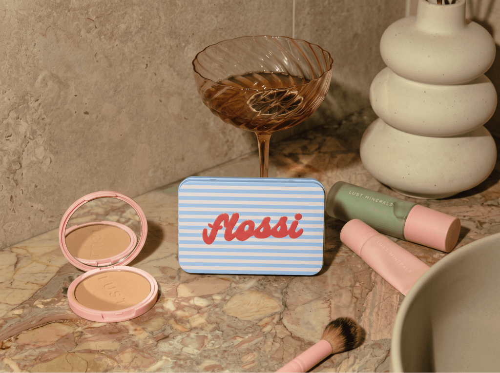 The striped blue Flossi travel case on a marble counter beside a compact and a cocktail glass